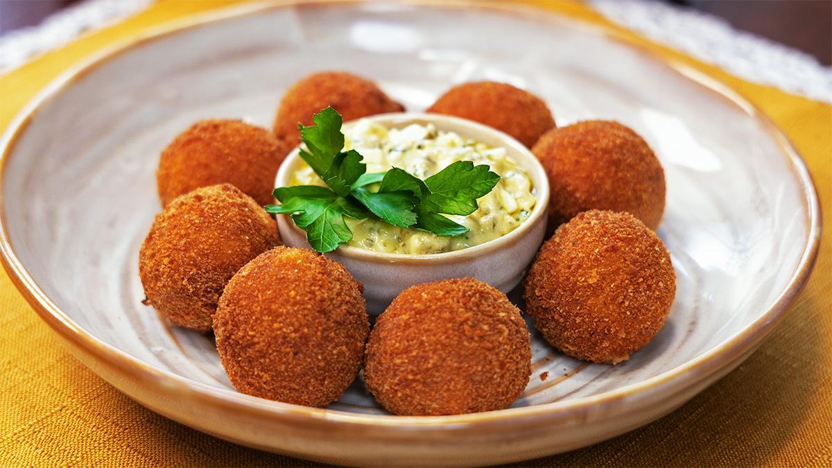 Croquettes with Gribiche Recipe This Croquettes with Gribiche Sauce recipe pairs crisp, golden croquettes with a tangy French sauce made from eggs, Dijon mustard, capers, cornichons, vinegar, fresh herbs, and oil. Featured on Dinner in Paris with host Romain Avril, these delectable French croquettes have a creamy béchamel-style filling made with cooked chicken or ham and a crunchy breadcrumb exterior. The Inspiration French croquettes are built around a simple idea: transform a creamy savoury filling into crisp, bite-sized portions. In this version from Dinner in Paris, host Romain Avril begins with a thick béchamel-style mixture of butter, flour, warm milk, and finely chopped chicken or ham. Chilling is essential to this French croquettes recipe. The mixture is spread onto a tray and refrigerated for one hour so it becomes firm enough to shape into one-inch balls. Each croquette is then coated in flour, beaten egg, and breadcrumbs before frying. The Gribiche sauce provides the ideal contrast. Cooked egg yolks are combined with Dijon mustard, white wine vinegar, salt, and pepper before olive oil and vegetable oil are slowly whisked in. Chopped egg whites, capers, cornichons, parsley, and tarragon are folded through at the end, creating a textured, tangy sauce that balances the richness of the fried croquettes. Ingredients For Croquettes 200 g (7 oz) cooked chicken or ham, finely chopped 40 g (3 tbsp) butter 1/4 cup flour 250 ml (1 cup) milk, warm ½ tsp salt ¼ tsp pepper Pinch of nutmeg 2 eggs, beaten 100 g (1 cup) breadcrumbs 50 g (½ cup) flour Vegetable oil, enough for 5 cm (2 inches) depth shallow frying For Gribiche Sauce 3 large eggs 1 tbsp Dijon mustard 2 tbsp capers, chopped 1/4 cup cornichons, chopped 2 tbsp white wine vinegar 1/4 cup extra virgin olive oil 1/4 cup vegetable oil 1 tbsp fresh parsley, chopped 1 tbsp fresh tarragon, chopped Salt and pepper, to taste Method Make Croquettes Melt butter in sauce pan over medium heat. Add flour and cook 2 minutes (do not brown). Slowly whisk in warm milk. Cook until thick, about 4-5 minutes. Stir in ham, salt, pepper and nutmeg. Spread mixture on tray and chill 1 hour until firm. Shape into 1 inch balls. Set up breading station with 3 bowls (eggs, flour and breadcrumbs separately). Dip each in the flour, then shake off excess. Place each ball in the egg, then let excess drip off. Finally, roll each ball in the breadcrumbs until fully coated. Place all the balls on a tray until ready to fry. Heat oil to 180°C / 350°F. Fry 3–4 minutes until golden brown and crispy. Let them sit for 1-2 minutes before serving. Make Gribiche Sauce Bring small pot of water to a boil. Place eggs in the boiling water gently and cook for 7.5 minutes. Remove the eggs from the water and place in an ice bath for 10 minutes. Once chilled, peel the eggs and cut them in half. Remove the yolks and place them in a mixing bowl. Chop the egg whites and reserve separately. To the bowl with the egg yolks, add dijon mustard, white wine vinegar, salt and pepper. Whisk until uniform. Ina small cup mix together the vegetable oil and olive oil. Slowly drizzle the oils into the egg yolk mixture and whisk vigorously. Once all the oil is in and the sauce is emulsified, fold in the egg whites, capers, cornichons, parsley and tarragon. Adjust seasoning and serve alongside the croquettes. Enjoy! Serving Suggestions These crispy French croquettes are ideal as an appetizer, small plate, or savoury snack. Arrange the golden croquettes on a platter with the Gribiche sauce in a small bowl on the side so each bite can be dipped into the tangy mixture of egg, Dijon mustard, capers, cornichons, and herbs. The sauce is especially important because its acidity cuts through the richness of the creamy filling and fried breadcrumb crust. The capers and cornichons provide briny sharpness, while parsley and tarragon bring freshness. Serve the croquettes shortly after frying so the exterior remains crisp while the inside stays soft and creamy. For entertaining, this French appetizer recipe works well as part of a larger small-plate menu. The croquettes are easy to portion individually, while the textured Gribiche sauce gives the platter a classic French character. Final Thoughts This Croquettes with Gribiche Sauce recipe combines crisp texture, creamy filling, and tangy French flavour in one satisfying appetizer. The croquette filling begins with a classic butter-and-flour base that thickens with warm milk into a smooth béchamel-style mixture. Find more recipes from Dinner in Paris here! Follow us on Instagram and YouTube for behind-the-scene footage and exclusive content!